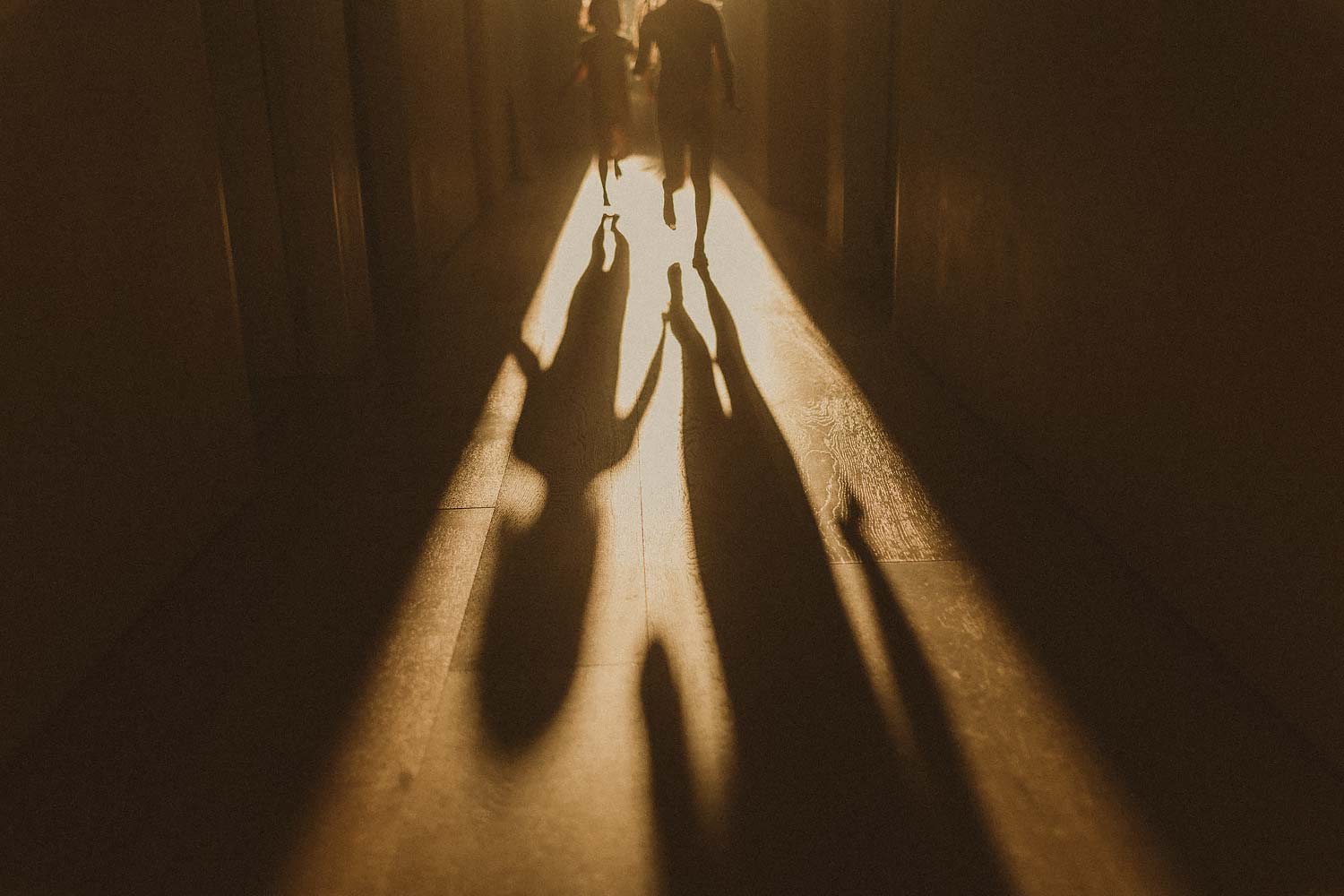 Family-photographer-sydney-two-children-run-playfully-down-a-hallway-towards-camera-casting-shadows-on-the-ground-from-the-sunlight-that-is-backlighting-the-image