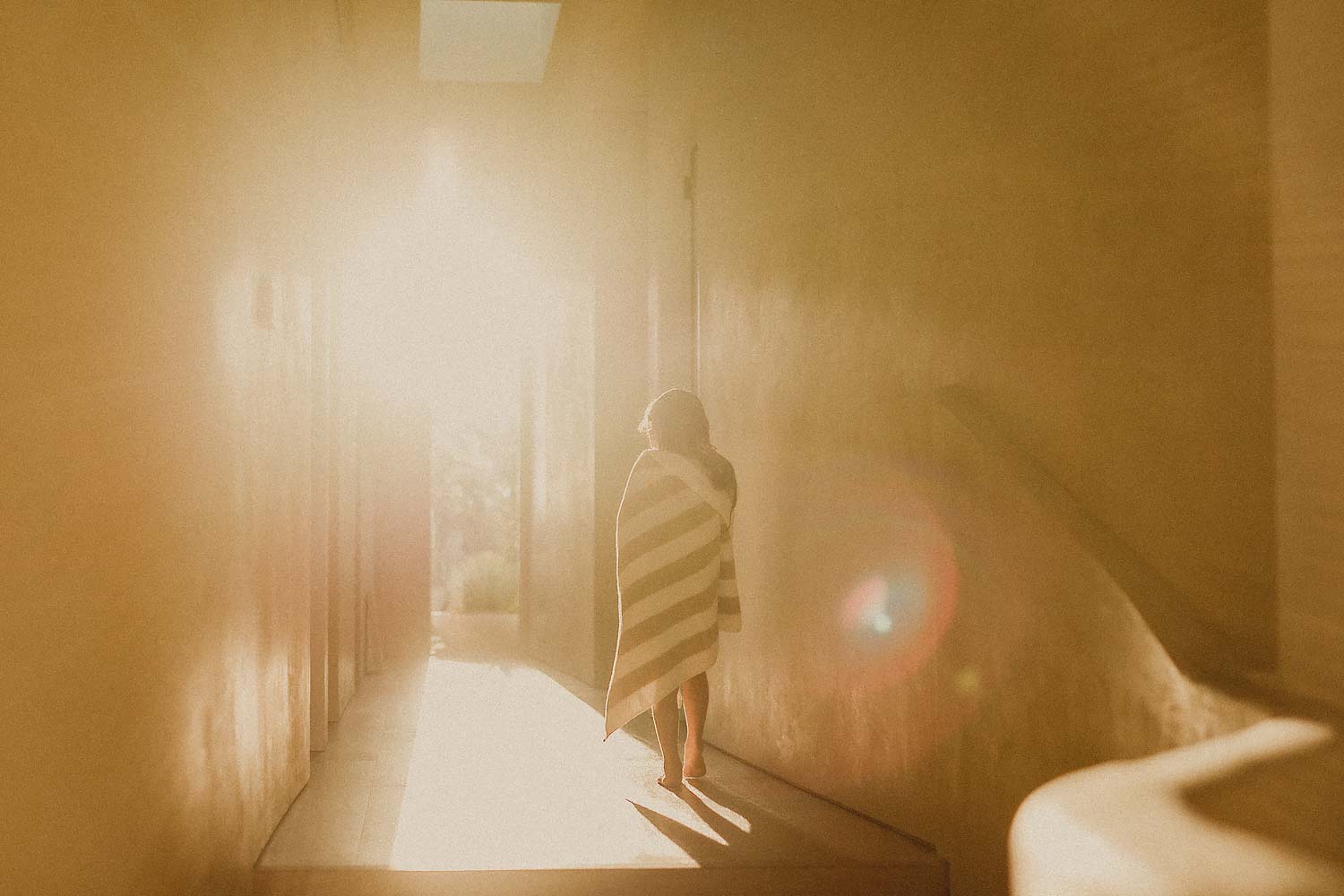 Family-photographer-sydney-young-child-facing-away-from-camera-walks-down-hallway-being-illuminated-by-golden-rays-from-the-sun-streaming-through-window-at-the-end-pf-the-hallway