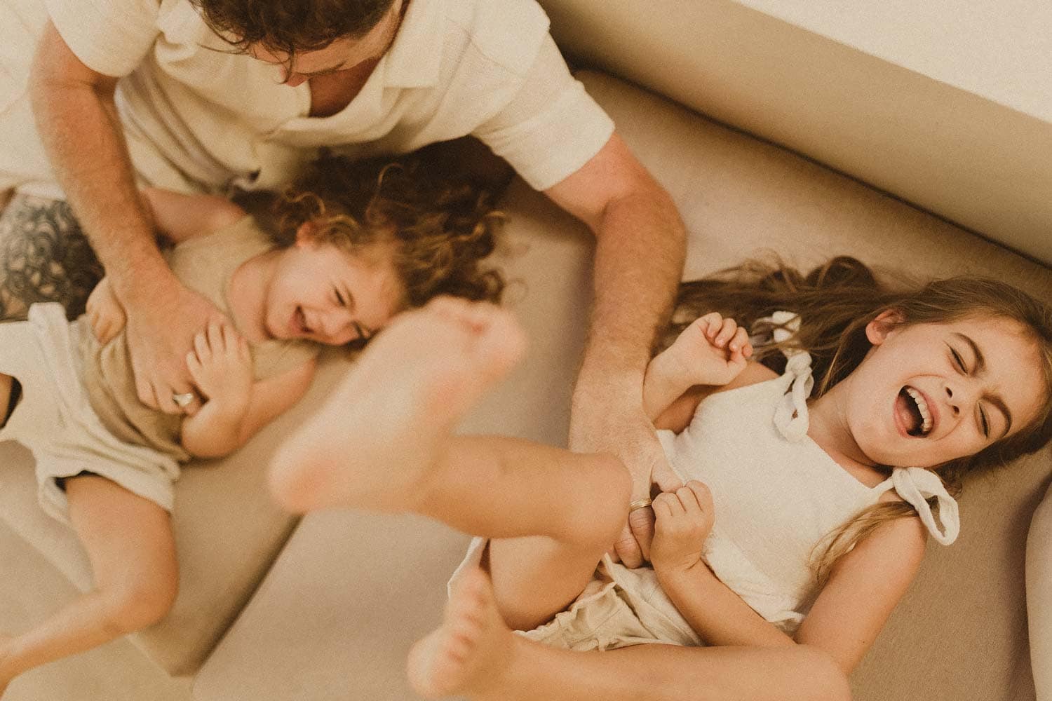 Family-photography-sutherland-shire-bireds-eye-view-of-young-girl-and-boy-being-tickled-on-a-beige-sofa-by-their-dad-whilst-they-giggle