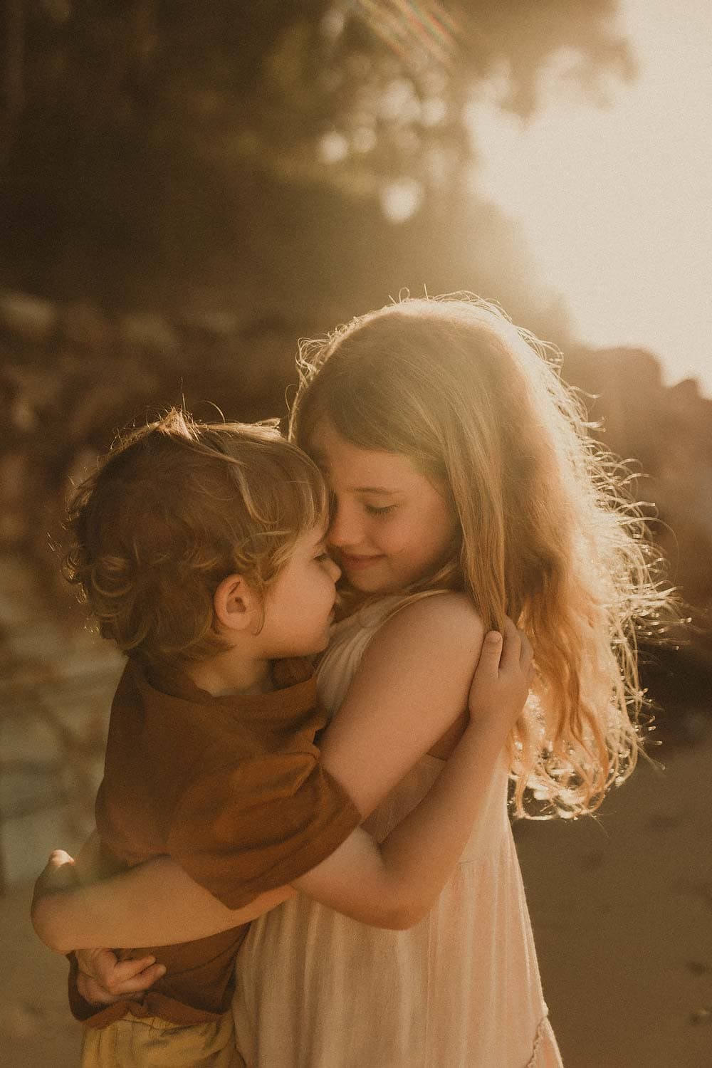Family-photography-sydney-little-boy-and-girl-embrace-sweetly-and-share-moment-with-little-girl-carrying-little-boy-and-little-boy-leaning-into-his-sister-whilst-the-sunlight-illuminates-her-hair-casting-golden-colours