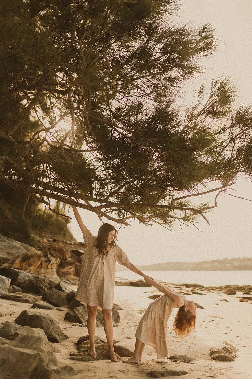 Lifestyle-Family-photographer-sutherland-shire-mum-holds-onto-tree-branch-that-overhangs-the-beach-and-rocks-whilst-holding-onto-hand-of-girl-who-playfully-leans-back-with-hair-free-flowing