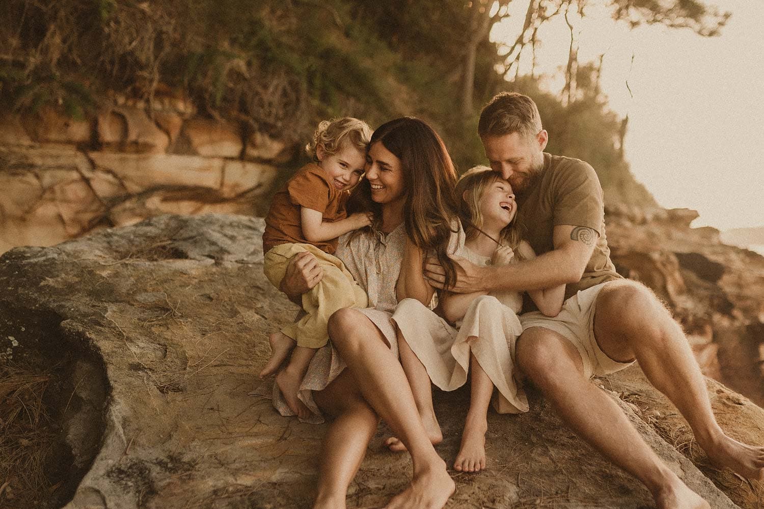 Lifestyle-family-photographer-sydney-mum-dad-young-boy-and-girl-sit-atop-a-rock-and-embrace-lovingly-whilst-laughing-with-dad-looking-at-daughter-as-she-laughs-and-mum-suggles-young-boy-as-he-leans-into-her-sweetly