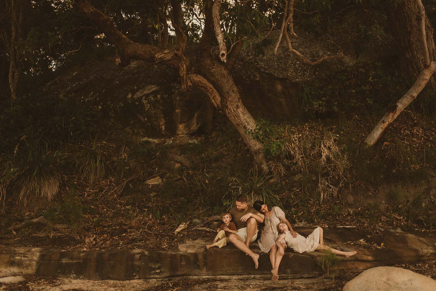 Sutherland-shire-family-photographer-family-of-four-sitting-on-a-large-flat-rock-amidst-rustic-bushland-background-mum-holds-onto-dads-arm-while-little-girl-rests-on-mum-and-looks-up-at-her-and-little-boy-sits-with-his-legs-tucked-under-next-to-his-dad-who-looks-down-at-him
