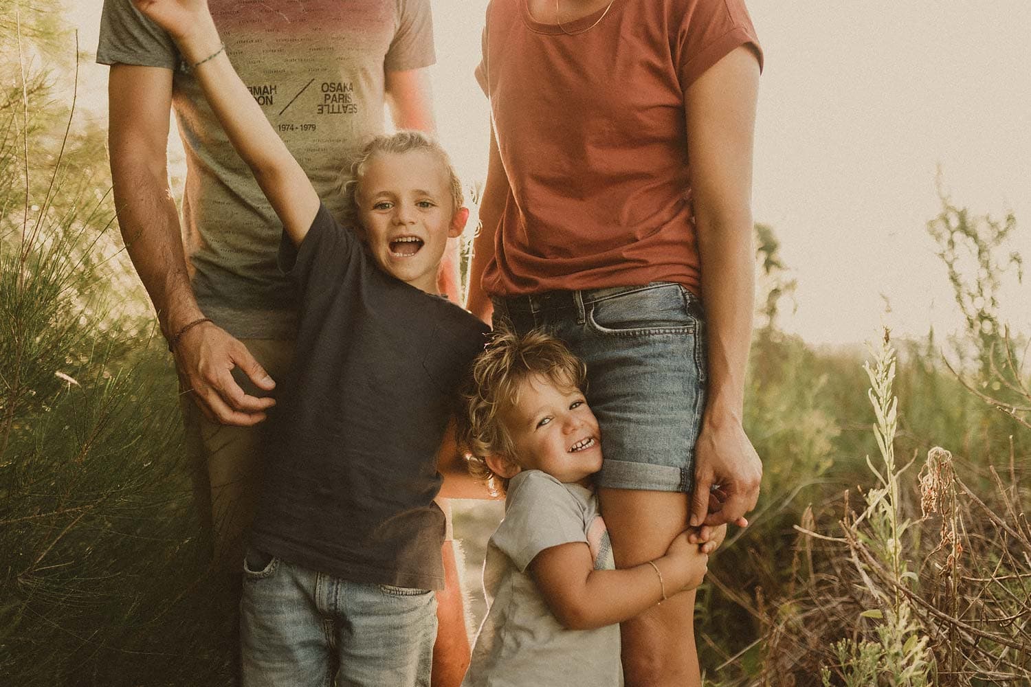 Sutherland-shire-family-photography-family-in-bushland-with-two-young-boys-smiling-at-camera-whilst-mum-and-dad-hold-them-in-background