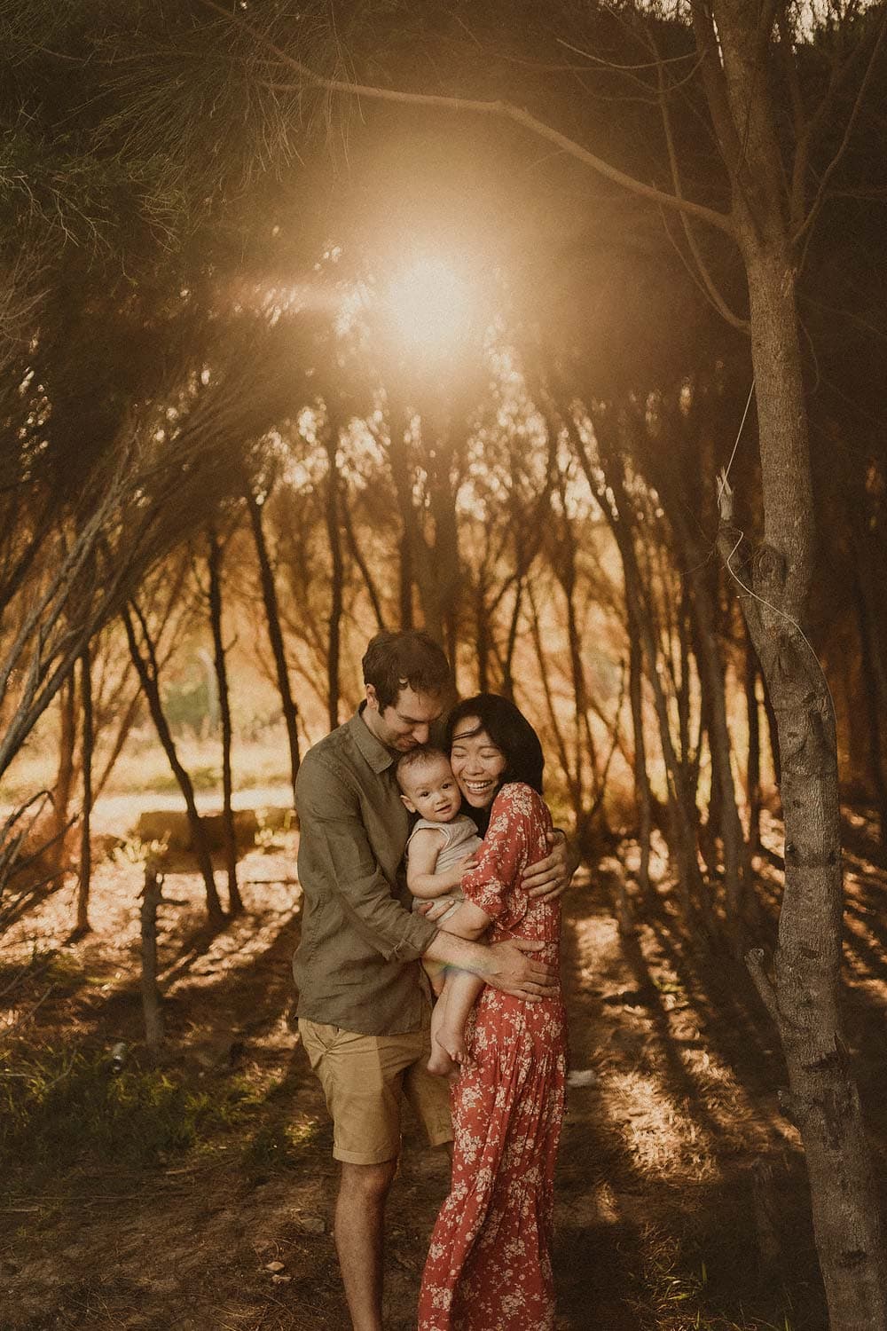 Sutherland-shire-family-photography-mum-and-dad-hugging-baby-with-golden-sunshine-streaming-through-trees-in-the-background