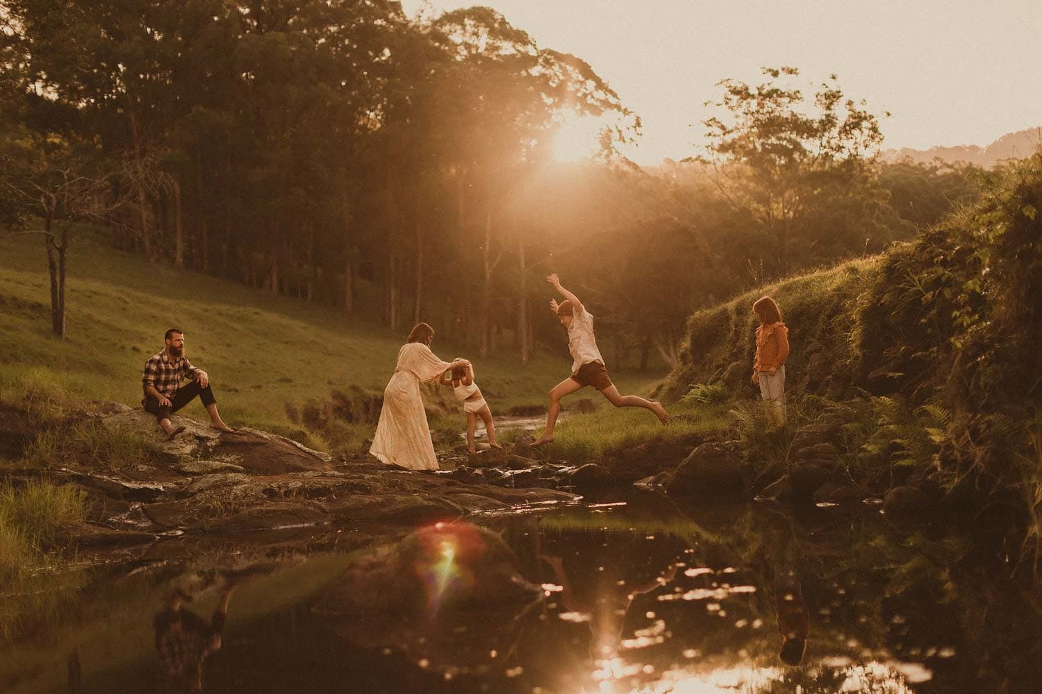 Sydney-family-photographer-sutherland-shire-family-jumping-rocks-in-river-with-boy-leaping-playfully-whilst-mum-helps-youngest-child-and-father-and-daughter-watch-on