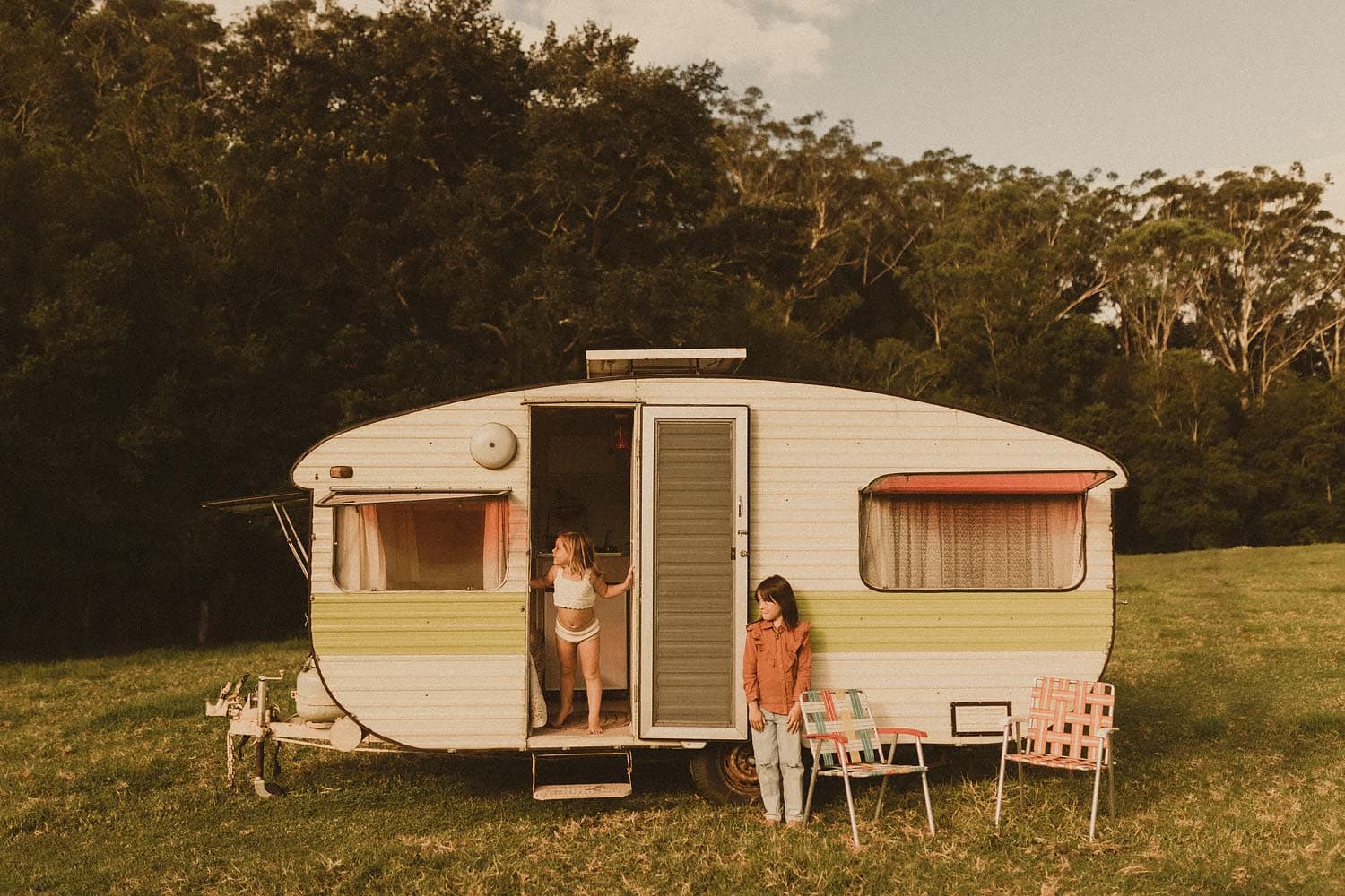 Sydney-inner-west-family-photographer-siblings-play-hide-and-seek-in-retro-caravan-set-in-grassy-meadow-with-vintage-deck-chairs