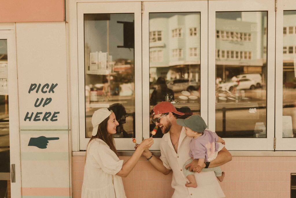 Family shoot at Bondi Beach Sydney, parents sharing rainbow icy poles with their baby girl outside a pastel cafe window in golden light
