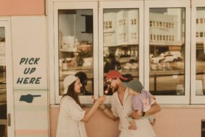 Family shoot at Bondi Beach Sydney, parents sharing rainbow icy poles with their baby girl outside a pastel cafe window in golden light