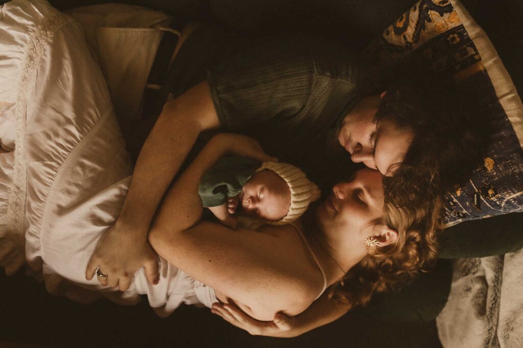 New dad kissing mum’s head as she cradles their sleeping newborn in a knitted bonnet, all three cuddled up in bed at home